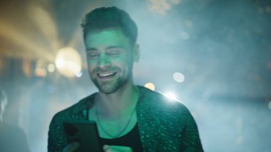 Karanlık gece kulübünde cep telefonunu izleyen hippi partileri. Gülümseyen adam parlak neon ışıklarla dans pistinde duran akıllı telefon mesajını okuyor. Disko kulübündeki mutlu adam gezici gezici uygulaması.