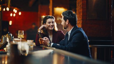 Gentleman lady relaxing together at night bar discussing smartphone news. Romantic pair scrolling phone enjoying soft lighting indoors. Happy couple exchanging words during evening gathering in pub
