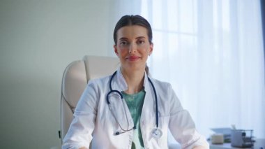 Gülümseyen doktor, klinik odası portresinde oturan kameraya bakıyor. Modern hastanede steteskopla poz veren başarılı bir kadın. İş molasında sağlık hizmetleri profesyonelliği hissi rahatladı