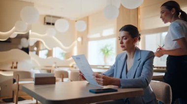 Modern restoranda menüyü inceleyen iş kadını garsonluk yapıyor. Zarif bayan kafeterya masasında tek başına yemek seçiyor. Profesyonel garson kız not defteri yazıyor. Müşteriden emir alıyor.