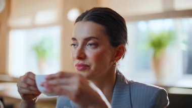 Modern şehir restoranında espresso içen bir işyeri sahibi. Gülümseyen kadın elinde sıcak içecekle kahvenin tadını çıkarıyor. Rahat kafe müşterisi kapuçino yudumlarken hafif restoranda mesafeye bakıyor