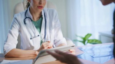 Hastane hekimi, sağlık sigortası ziyaretinde hastayla konuşuyor. Beyaz önlüklü yetenekli gülümseyen doktor tablet bilgisayarlı ekranda bayan ziyaretçiyle sağlık tedavisini tartışıyor. Sıhhiyeci kadın çalışıyor