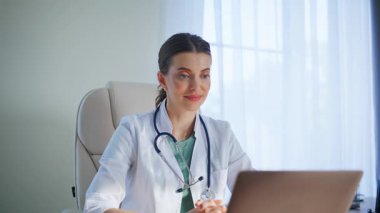 Sıhhiyeci klinikteki bilgisayarlı ekranda çevrimiçi danışmadan bahsediyor. Steteskopla gülümseyen tıbbi uzman video sağlık hizmetlerine odaklanmış durumda. Doktor teletıp işiyle uğraşıyor.