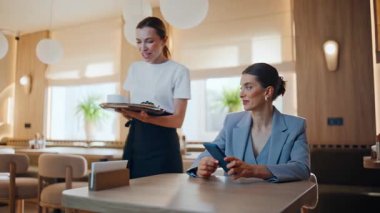 Gülümseyen kadın kafeterya masasında oturmuş rüya gibi bir yüz ifadesi olan akıllı telefon kullanıyor. Dost canlısı restoran garsonu bayan müşteriye kahve fincanı veriyor. Keyifli bayan rahat kafeteryada yalnız başına dinleniyor.