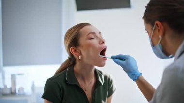 Sıhhiye klinik muayene odasındaki hastanın boğazını inceliyor. Sağlık teşhisine odaklanmış özel ekipman kullanan bir tıp uzmanı. Kadın poliklinik ziyaretçisi klinik kontrolü için ağzını açıyor.