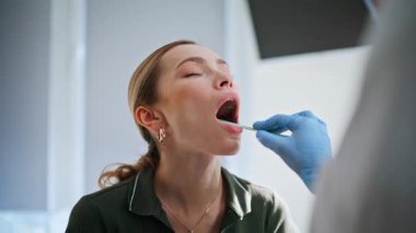 Klinikteki hasta sağlık kontrolünde ağzını açar. Kadın pratisyen hekim poliklinik ziyaretçisinin sözlü muayenesini yapıyor. Koruyucu eldivenler konusunda uzman. Kadın boğazına dikkatlice bakıyor.