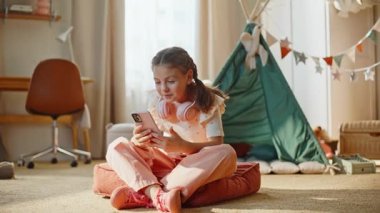 Konforlu oda halısında akıllı telefon oynayan bir çocuk. Küçük bir çocuk cep telefonuyla sabah apartmanında internette geziniyor. Heyecanlı küçük kız cep telefonuyla ilgileniyor.