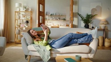 Cep telefonuyla rahat kanepe kullanan yalancı bir kadın. Gülümseyen çekici kız, video aramasından zevk alıyor. Güneş ışığı dairesinde selfie çekiyor. Kaygısız neşeli bayan koltukta yalnız başına cep telefonuyla dinleniyor.