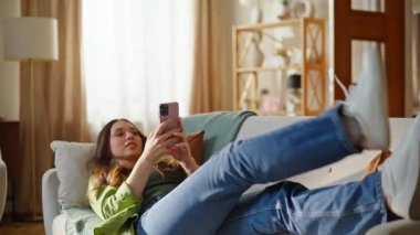 Akıllı telefon ekranı seyreden rahat bir kız rahat koltukta uzanıyor. Gülümseyen genç kadın cep telefonu dinlendirici kanepesinde haber yayını yapıyor. Güneş ışığı apartmanındaki cep telefonunda gezinen mutlu bayan