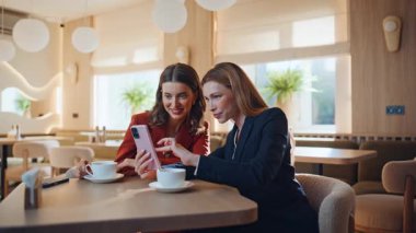 İş kadınları öğle yemeğinde modern restoranda oturarak akıllı telefon izliyor. Kafede internetteki telefon içeriğiyle gülümseyen iki kadın. Kahve molasında cep telefonuna bakan mutlu iş kadınları.