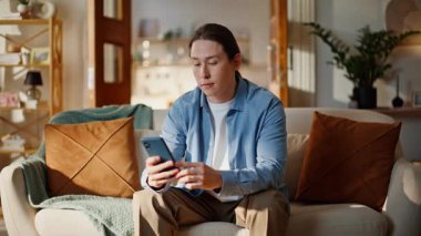 Bej koltukta oturmuş cep telefonuyla sohbet eden sakin bir adam. Rahatlamış esmer, kanepede dinleniyor. Oturma odasında sosyal medya okuyor. İçeride dinlenen mutlu gülen adam. Teknoloji ve insanlar kavramı