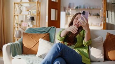 Web kamerasında konuşan gülümseyen kadın akıllı telefondan koltuğa yakın oturarak sohbet ediyor. Güzel bayan evde cep telefonuyla hayat haberlerini tartışıyor. Kız, erkek arkadaşıyla cep telefonuyla konuşuyor.