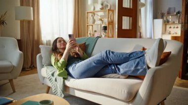 Yalancı kız akıllı telefondan mesaj atıyor rahat kanepe yastıkları dinleniyor. Evde cep telefonu okurken gülümseyen mutlu esmer kadın. Neşeli kadın cep telefonlarını karıştırıyor sosyal medya rahatlatıcı kanepe