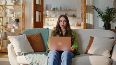 Konforlu apartman kanepesinde oturmuş dizüstü bilgisayarla internette sörf yapan kadın. Hafif modern oturma odasındaki cihazda yorgun esmer yazıcı e-postası var. Çevrimiçi işi bitiren yorgun kadın bilgisayarın kapağını kapalı tutuyor