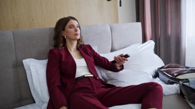 Lüks otel odasında televizyon seyreden gülümseyen bir gezgin. Uzaktan kumandalı yatağı kullanarak kanal seçen mutlu iş kadını. Beyaz yataklarda dinlenen pozitif kadın televizyonun tadını çıkarıyor..