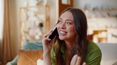 Rahat oturma odasında cep telefonuyla konuşan kaygısız bir kadın. Rahatlamış genç bayan sabahın erken saatlerinde akıllı telefonu arıyor. Kanepede otururken telefonla konuşan nazik bir kız. Dostça iletişim.