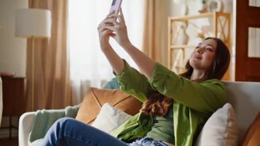 Bayan blogger modern oturma odasında video içeriği kaydediyor. Mutlu neşeli kız abonelerle cep telefonu kullanarak iletişim kuruyor. Kanepede akıllı telefondan olumlu kadın etkisi yayılıyor.