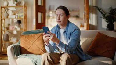 Kanepeye yakın çekimde akıllı telefon yazan Asyalı adam. Rahat iç mekanda cep telefonuyla dinlenen pozitif rahatlamış ssm işçisi. Sakin bir insan gündüz vakti koltukta tek başına dinleniyor.