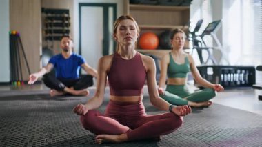 Nilüfer pozisyonunda oturan kapalı gözlü insanlar modern spor salonunda farkındalık alıştırması yapıyorlar. Spor kulübünde yoga sınıfıyla meditasyon yapan barışçıl kadın antrenör. Üç dingin yoga ustası beden ve zihin dengesi kuruyor..