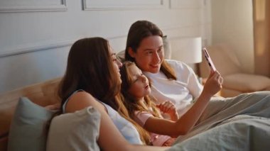 Akıllı telefon kullanan neşeli bir aile güneş ışığı yatak odasında fotoğraf çekiyor. Modern cep telefonu kamerasına poz veren çocuklu mutlu çift yumuşak yatakta oturuyor. Neşeli ebeveynler küçük çocukla eğleniyor.