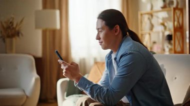 Esmer, geveze, cep telefonuyla sohbet eden bej koltuğa yaslanmış, yakın plan. Rahat adam oturma odasında sosyal medya okuyarak kanepede dinleniyor. İçeride dinlenen mutlu gülen adam. Teknoloji ve insanlar kavramı