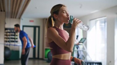 Sporcu kadın spor salonundan sonra dinlenmek için su içiyor. Spor giyim bölümündeki zinde kadın ayakta duran şişeden taze sıvı yudumluyor. Spor antrenmanında ince esmer ferahlatıcı. Sağlıklı vücut.