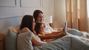 Pozitif aile tablet bilgisayarı tutuyor. Yatakta yan yana yatıyorlar. Çocuk kızıyla gülümseyen ebeveynler içerideki modern cihazlarda komik videolar izliyorlar. Mutlu akrabalar rahat bir dairede sekme kullanıyor.