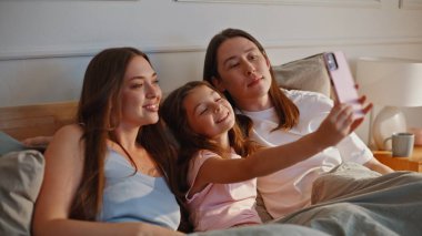 Pozitif aile, güneş ışığı yatak odasında akıllı telefon kullanarak fotoğraf çekiyor. Modern cep telefonu kamerasına poz veren çocuklu mutlu ebeveynler yumuşak yatakta oturuyor. Neşeli anne, baba ve çocuk eğleniyor.