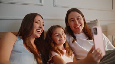 Neşeli aile, rahat yatak odasında cep telefonuyla selfie çekiyor. Yavru kızı olan mutlu çift, yumuşak yastıkların üzerinde yalan söylüyor. Çocuklu neşeli ebeveynler akıllı telefona el sallıyor.