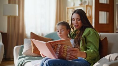 Modern evde masal okuyan sevimli bir anne çocuğu. Oturma odasında yakın plan. Mutlu sevgi dolu aile hafta sonu apartmanında sohbet ediyor. Küçük kız anne babasıyla konuşuyor rahat koltukta oturuyor.