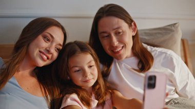 Ebeveyn kızı güneş ışığı yatak odasında selfie çekiyor. Mutlu anne baba, evde akıllı telefon kamerasına bakan küçük kızla gülümsüyor. Tasasız anne baba, küçük çocuk aile fotoğrafı çekiyor.