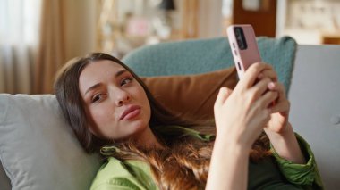 Modern apartmandaki cep telefonundan mesaj atan rahat bir kız. Gülümseyen hayalperest kadın evde sosyal medya akıllı telefonlarına bakıyor. Neşeli bayan internetten alışveriş yapıyor oturma odasında cep telefonu kullanıyor.