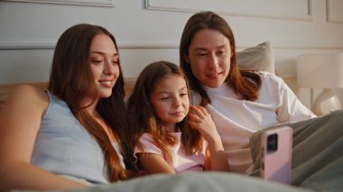 Mutlu bir ailenin Sunny House 'da videolu yayını var. Küçük çocuklu gülümseyen genç ebeveynler sabah yatak odasında cep telefonu kullanıyor. Küçük kızıyla neşeli bir çift cep telefonuyla konuşuyor.