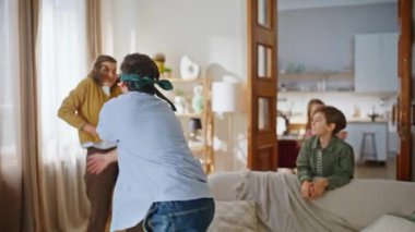 Oyuncu baba, anneyi gözleri bağlı oturma odasını kapatırken yakalıyor. Gülen ebeveynler, saklambaç oynayan sevimli çocuklar aile içi oyun konforlu evlerinde vakit geçirirler. Gülümseyen sakallı adam karısına sarılıyor.