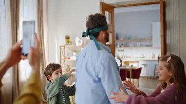 Gözü bağlı babaları dönen komik çocuklar yakın çekimde eğleniyorlar. Ebeveyn çocukları hafta sonu oyunu oynuyor. Çocukları yakalamaya çalışan gözleri bağlı bir adam. Akıllı telefon çeken bir kadın. Mutlu aile eğleniyor.