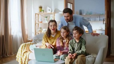 Neşeli ebeveynler laptopla ev koltuğunda otururken görüntülü arıyorlar. Gülümseyen sakallı baba güzel kadın reşit olmayan çocuklar sanal buluşmada konuşuyor web kamerasına bakarak sohbetin tadını çıkarıyor.