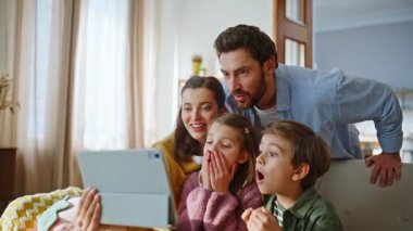 Modern apartmandaki web kamerasına işaret eden tabletle neşeli aile videosu. Şaşırmış çocuklar bilgisayarlı web kamerasıyla konuşuyor. İnternette ailelerle oturup eğleniyorlar.