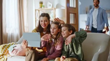 Anne çocuklar video çağrısını tablet konforlu kanepe ile karşılıyorlar. Sakallı baba sanal sohbete katılıyor. Neşeli bir aile içeride el sallıyor. Uzak sohbet kavramı