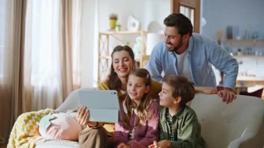 Anne ve babaları tablet bilgisayarı hazırlayan video oyunu oynuyorlar. Gülümseyen neşeli akrabalar el sallıyor. Sevdikleriyle sanal olarak buluşuyorlar. Parlak, düz oturma odasında internet kullanıyorlar.