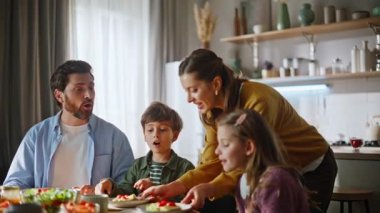 Şefkatli anne kahvaltı sofrasını hazırlıyor. Gülümseyen anne tabakları koyuyor ve çocukları öpüyor. Dost canlısı insanlar hafta sonunu rahat bir dairede geçirirler. Ebeveynler çocuklar sabah evde.