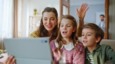 Anne tableti yakın çekim tutuyor. Gülümseyen akrabalar aydınlık oturma odasındaki modern aletlere video çağrısında bulunuyorlar. Mutlu ebeveynler, çocuklar el sallayarak bilgisayar ekranına bakıyorlar. Neşeli aile blogları