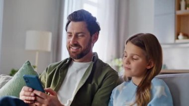 Tasasız bir aile, hafta sonu rahatlamış ev çekimlerinde dijital eğlencenin tadını çıkarıyor. Gülümseyen baba sevimli görünümlü genç kız akıllı telefona göz atıyor sosyal medya internette geziniyor..