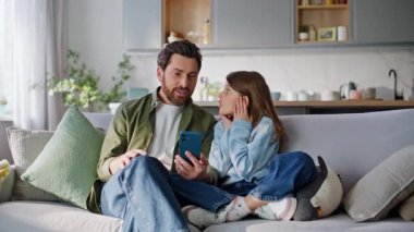 Baba kız cep telefonuyla sosyal medya içeriğini tartışıyor birlikte rahat bir daire kanepesinde oturuyor. Mutlu sakallı adam, genç kız hakkında konuşuyor. Akıllı telefon görünümlü, çevrimiçi uygulama kapalı alanda geziniyor..