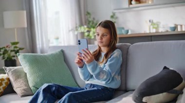 Tasasız genç bayan cep telefonunu karıştırıyor rahat kanepeyi tek başına dinlendiriyor. Gülümseyen genç kız internette rahat bir apartman kanepesinde oturup sosyal medyayı izlemekten zevk alıyor. Çocuklar telefon bağımlılığı