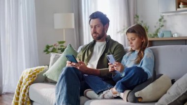 Rahat aile telefonları rahat bir kanepede yan yana. Gülümseyen sakallı baba akıllı telefon seyrediyor. Modern evde oturan sevimli genç kız gibi görünüyor. Dijital bağımlılık.