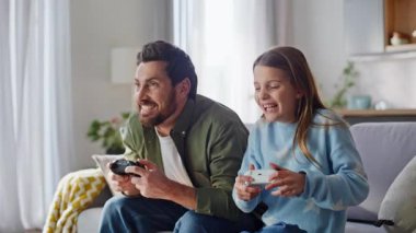 Duygusal kız baba online oyun oynuyor. Evdeki kanepeye yakın oturuyor. Rekabetçi baba çocuğu beş kez kucaklaşarak oyun kazanıyor birlikte eğleniyoruz. Gülümseyen adam elinde joystickle.