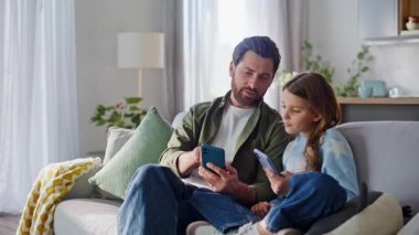 Baba kız rahatlamış aile hafta sonlarında oturma odasındaki koltukta telefonlarını karıştırıyor. Gülümseyen sakallı baba, cep telefonuyla kızının rahat koltuğunu gösteriyor. Mutlu modern aile dijital hayata dahil..