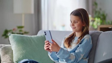 Genç kız akıllı telefon görünümlü rahat ev koltuğu yakın çekim. Gülücükler saçan, sosyal medyada cep telefonuna bakan güzel bir genç kız. Rahatça evde oturuyor. Genç yaşam tarzında modern teknoloji.