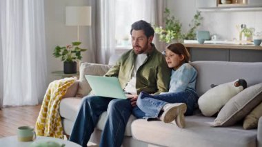 Modern dairede el sallayan çocuk videosu tableti arıyor. Gülümseyen baba bilgisayarlı web kamerasıyla konuşuyor. Ev kanepede rahat rahat oturarak eğleniyorlar. Aile teknolojisi yaşam tarzı.