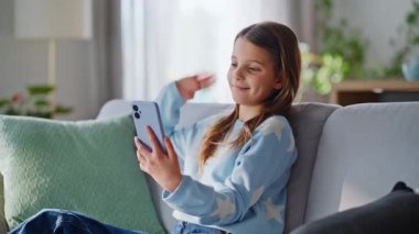 Genç kız cep telefonunu modern oturma odasındaki kanepeye yakın çekiyor. Gülümseyen ergen akıllı telefonlu webcam görüntülü görüşme sanal dersini evde oturarak alıyor. Modern dijital yaşam.